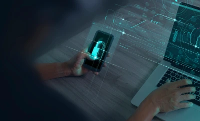 Person holds a smartphone displaying a glowing digital lock while a laptop shows a security dashboard, suggesting cybersecurity activity.
